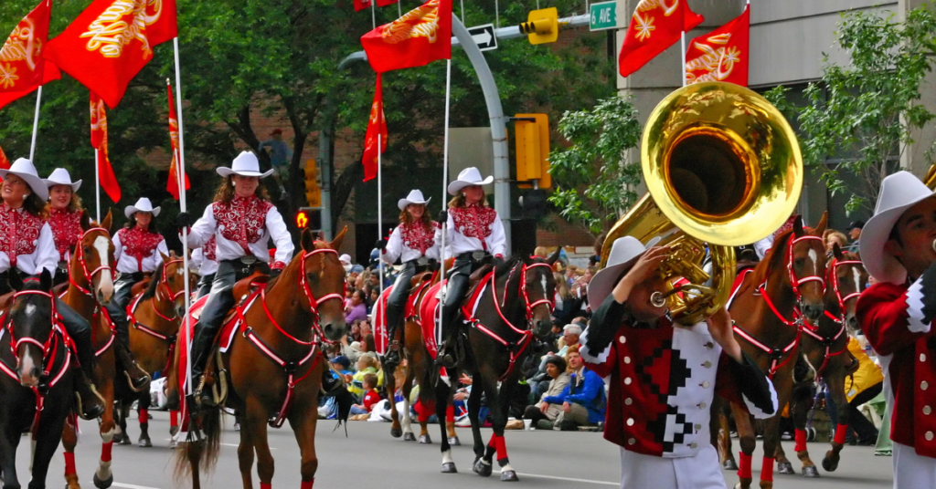 Vibrant street parade scene with horseback riders carrying red and gold flags and marching band members in red and white uniforms at the Calgary Stampede, reflecting the festive spirit and cultural heritage of this renowned rodeo event.
