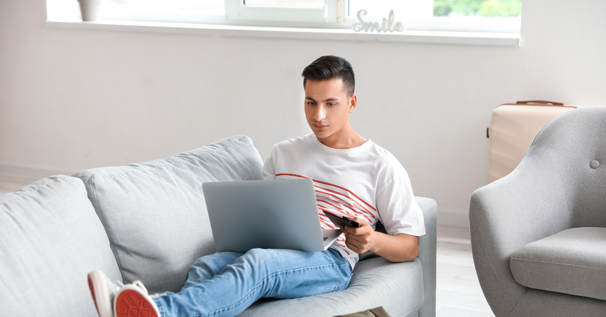 Homme recherchant des offres de vol sur un ordinateur portable alors qu'il est assis sur un canapé à la maison, illustrant la réservation de voyages en ligne intelligente.