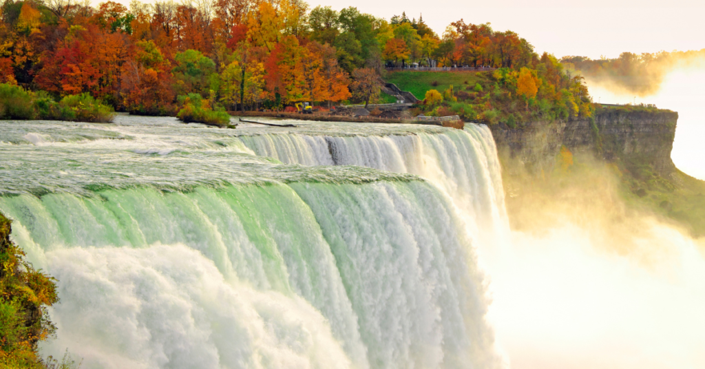 Vivid image of Niagara Falls during autumn with mist rising from the cascading waters and trees displaying vibrant fall colors in the background, showcasing the natural beauty and power of this famous landmark.